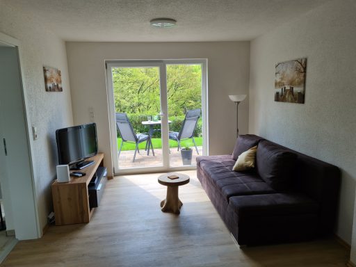 Helles Wohnzimmer mit Sofa, Fernseher und Blick auf eine Terrasse mit Stühlen.
